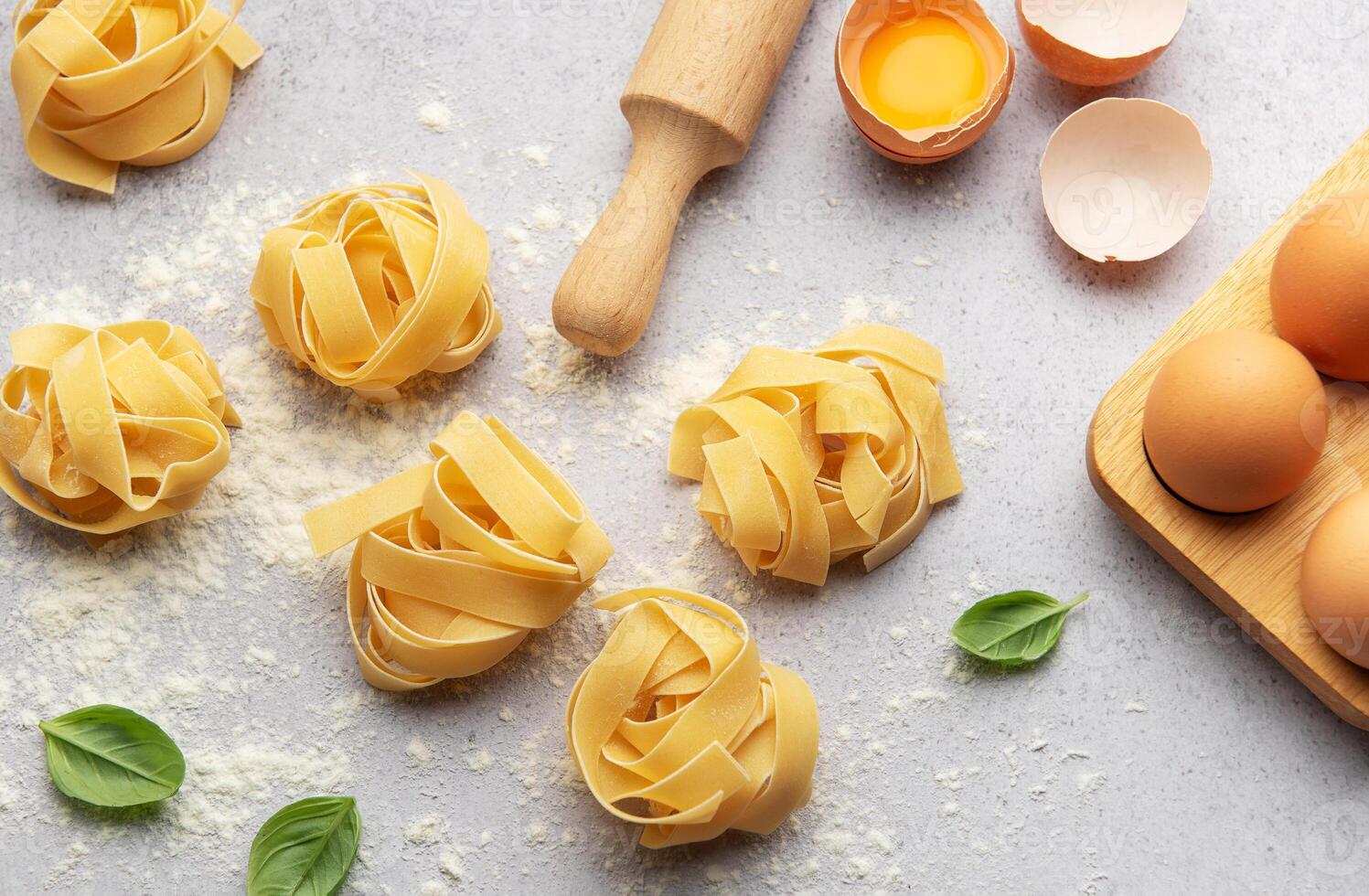 Fettuccine pasta nests with basil, eggs, flour and rolling pin on kitchen table photo