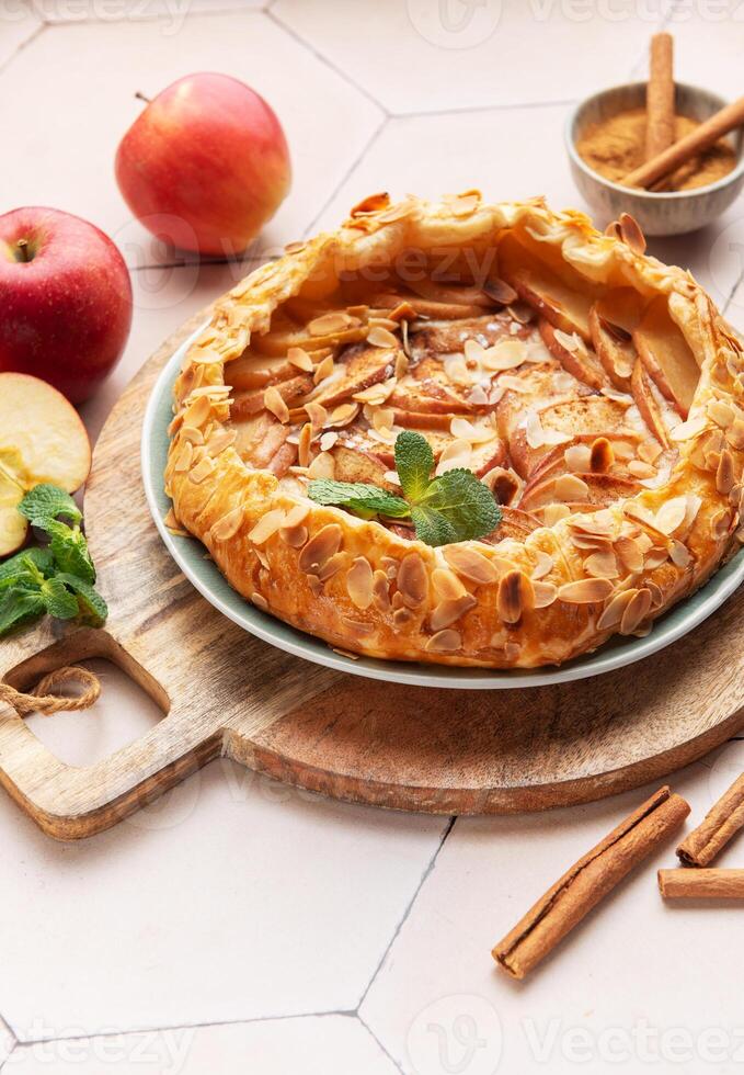 Homemade apple tart with almond toppings on a wooden board surrounded by fresh apples and cinnamon photo