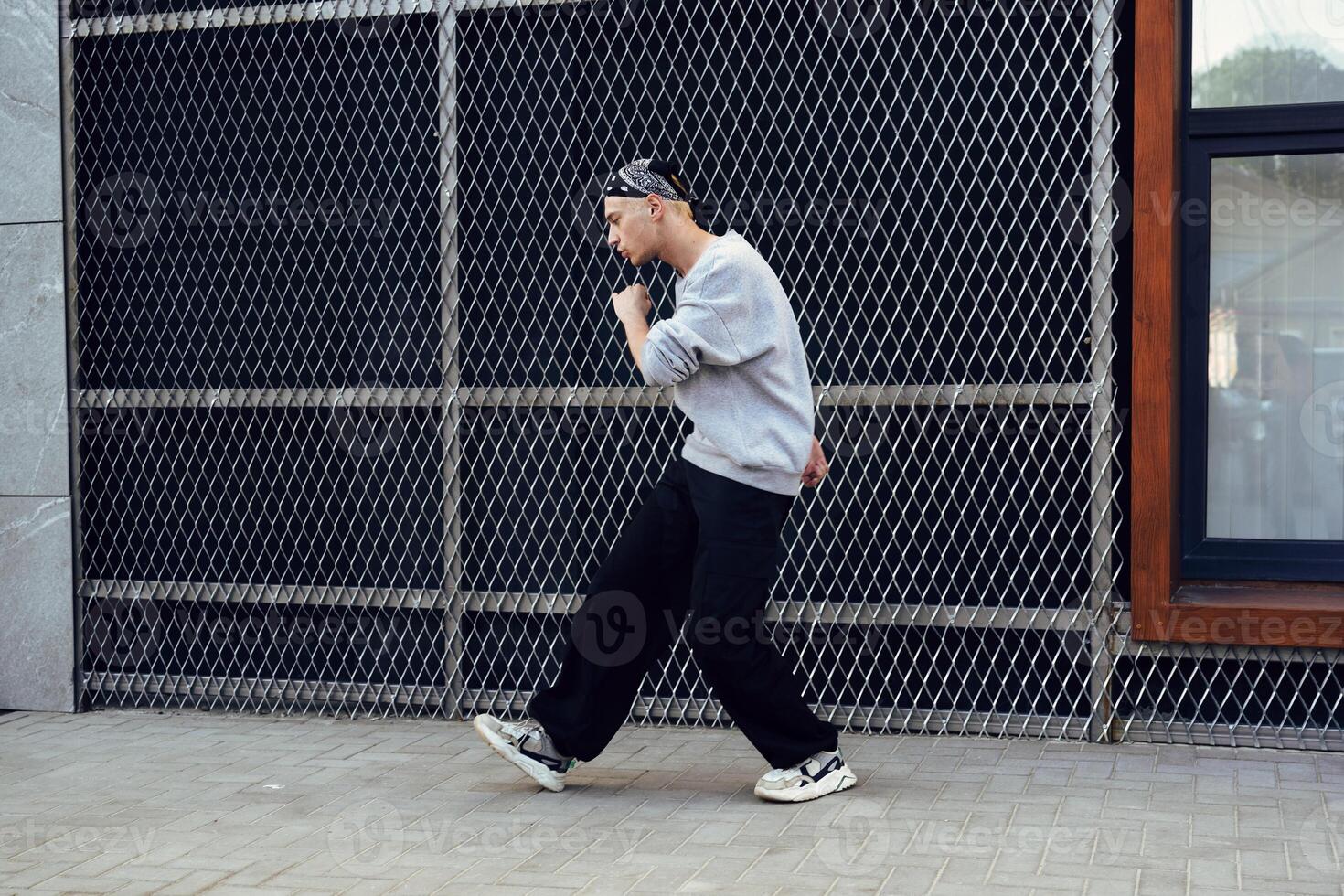 A man wearing a gray sweater and black pants dances in front of a chain-link fence in the daytime photo