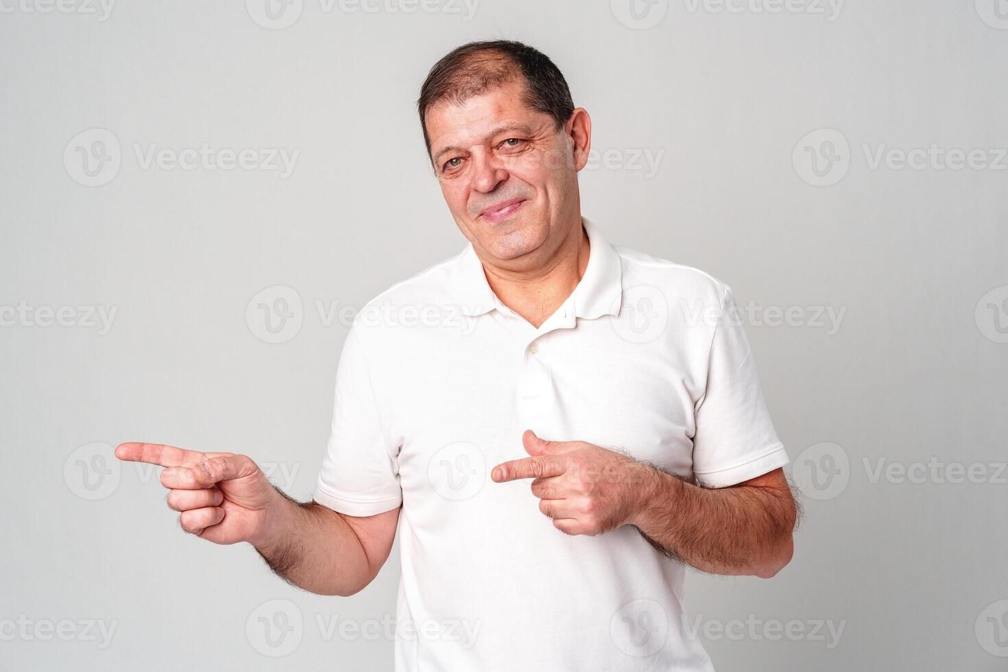 Man dressed in a white polo shirt and jeans happily pointing to the side against a plain background photo