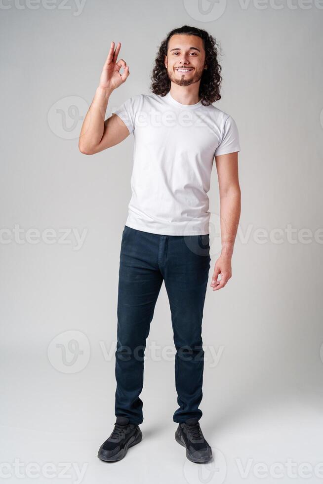 A young man in a white t-shirt and jeans confidently shows an okay hand gesture photo