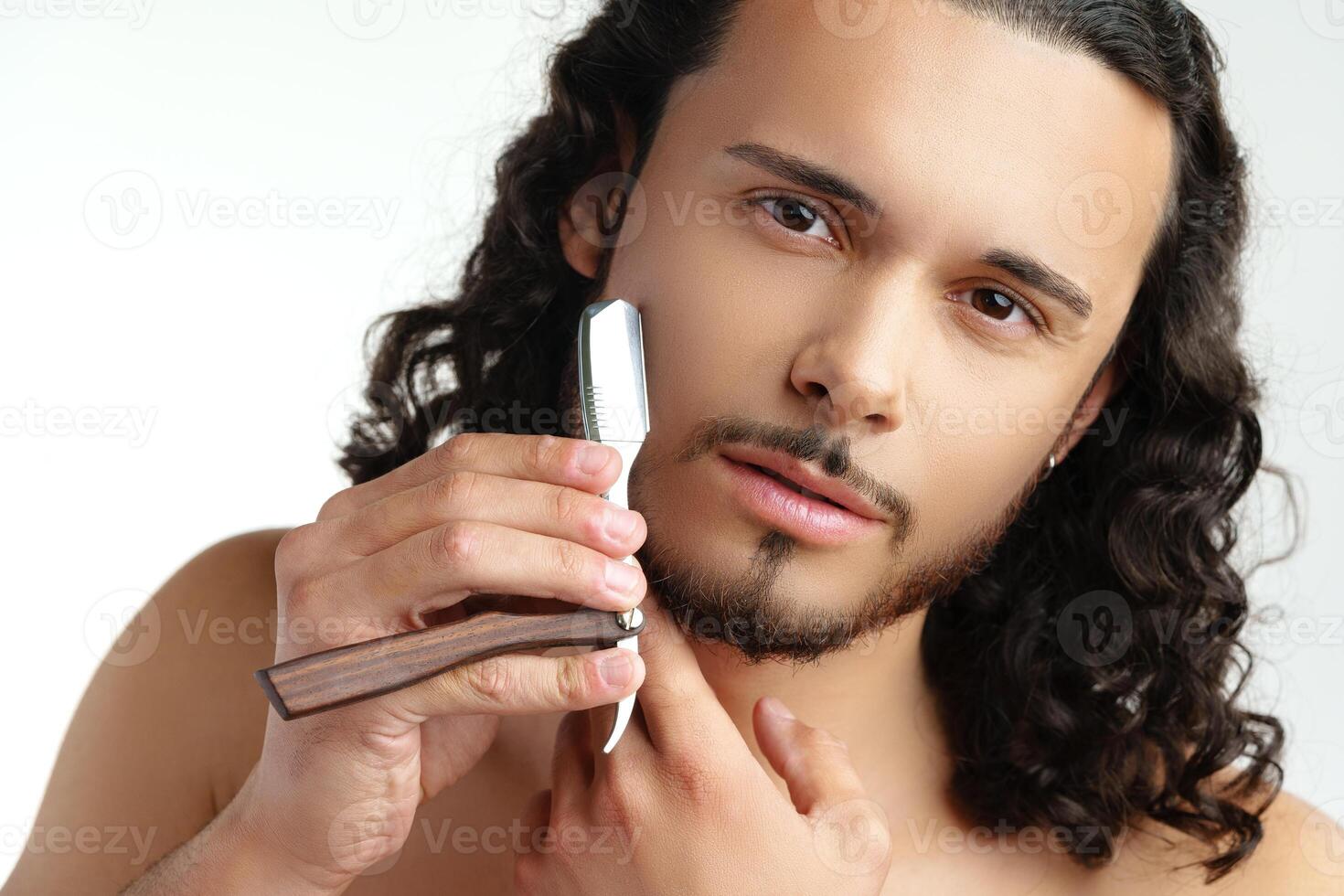Man with curly hair grooming his beard carefully using a straight razor against a plain white background photo