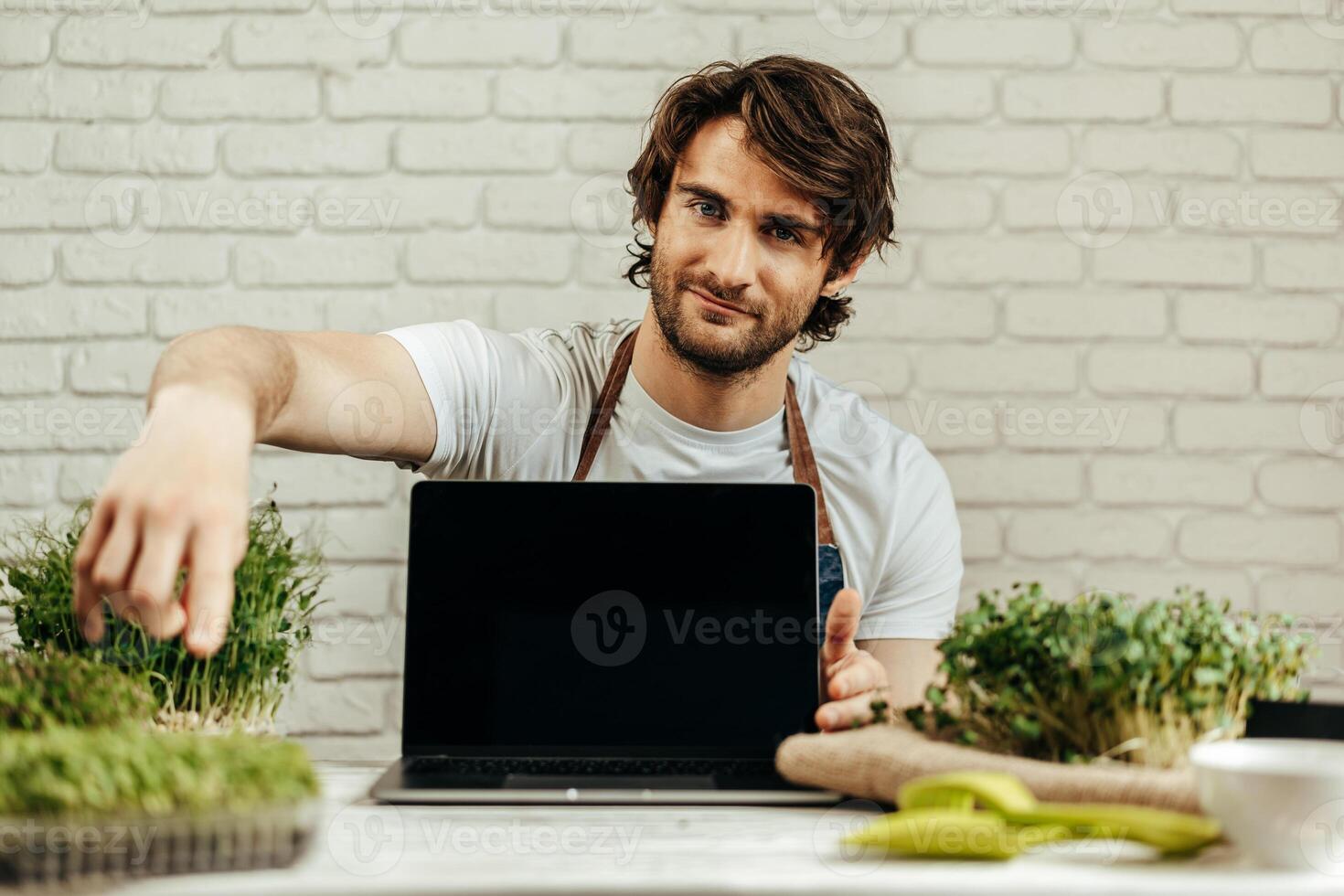 Man farmer shows black screen of laptop and sits at the table with sprouts photo