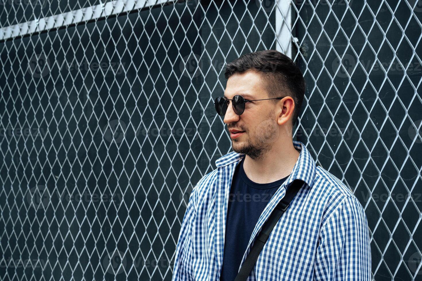 A man in a blue checkered shirt smiles while standing outside a modern building on a sunny day photo