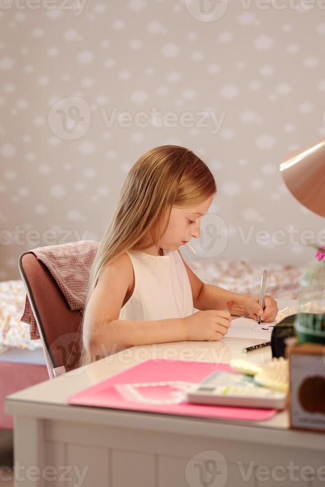 A young girl with long blonde hair sits at a desk in a room with a cloud patterned wall, concentrating on drawing in her notebook photo