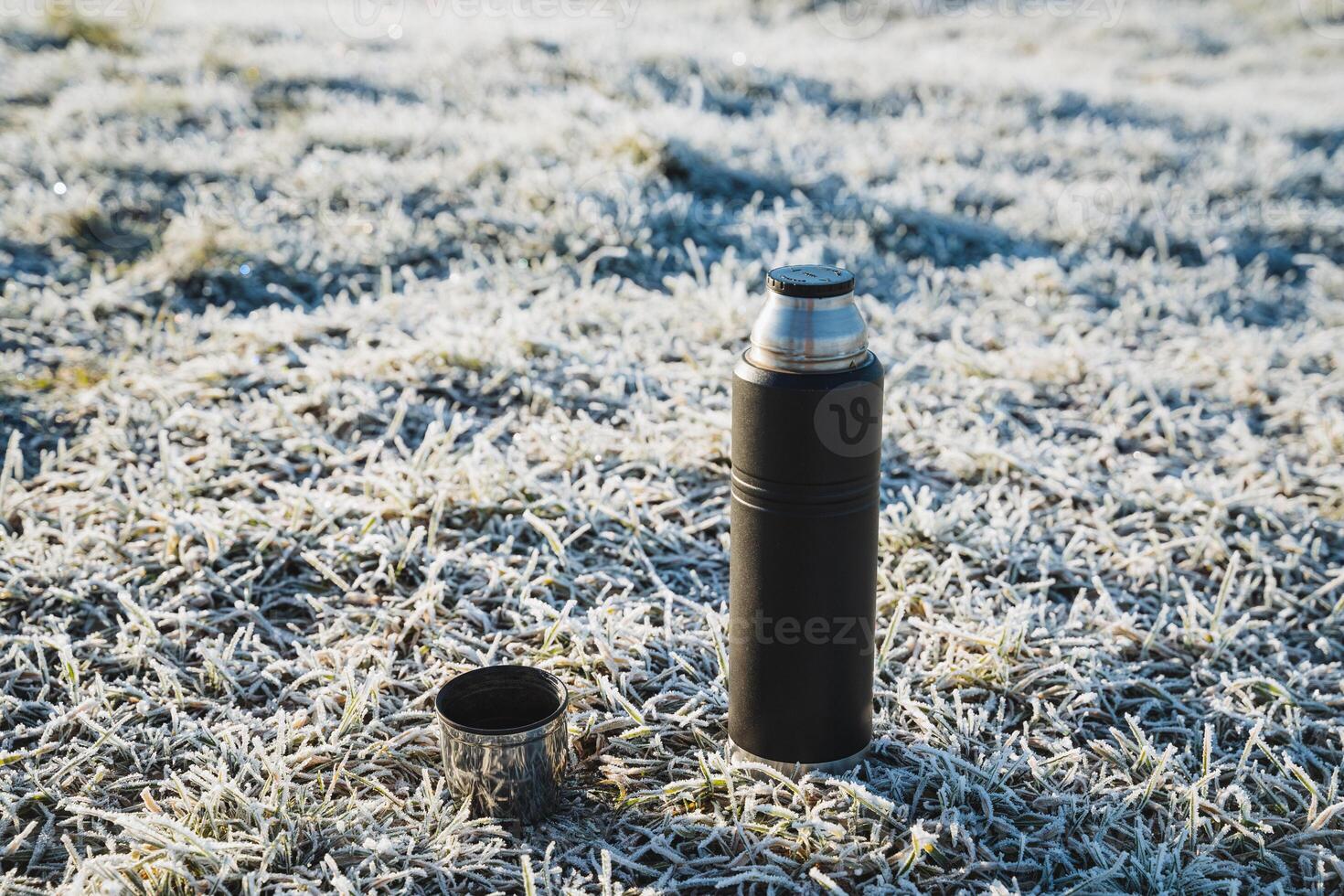 Thermos standing on the ground in cold weather, tea mug on a hike, camping utensils, hot drink in a metal flask. photo