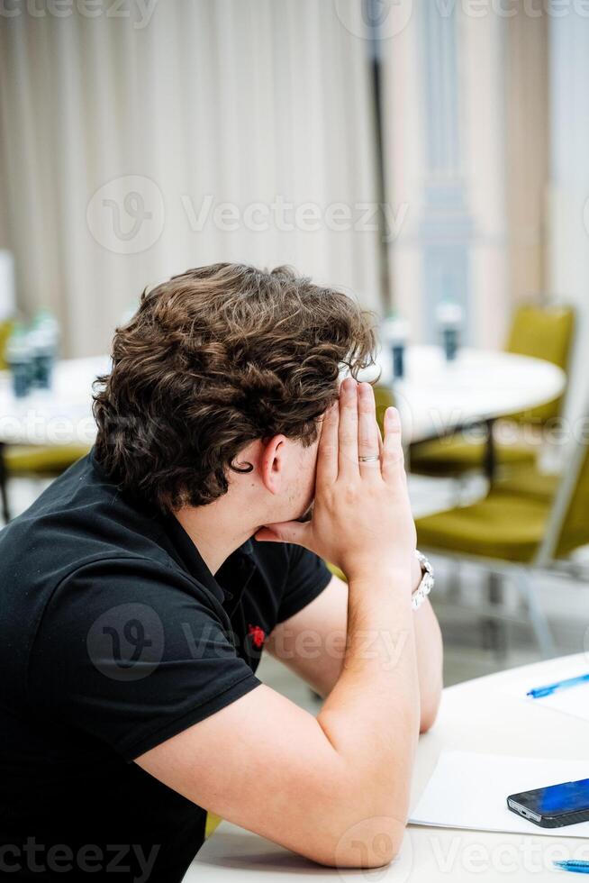 A man is sitting at a desk with his hands covering his face, a man is sitting in an office. The young man turned his head to the side. photo