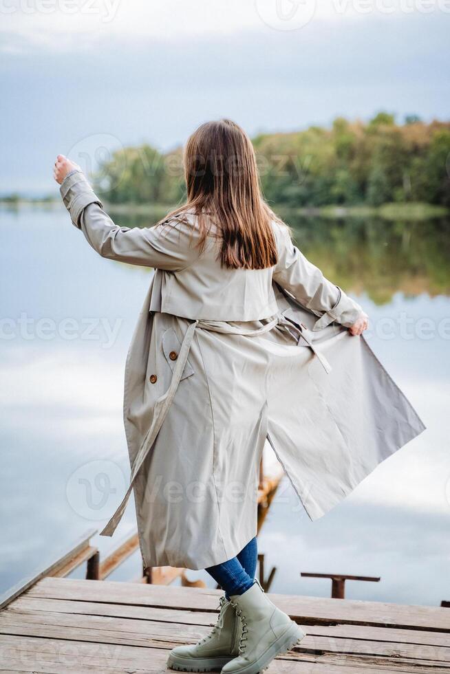 Girl dancing on the bridge in a raincoat, gray casual style raincoat, long hair at the back, loose curls, good mood, walk on the lake. photo