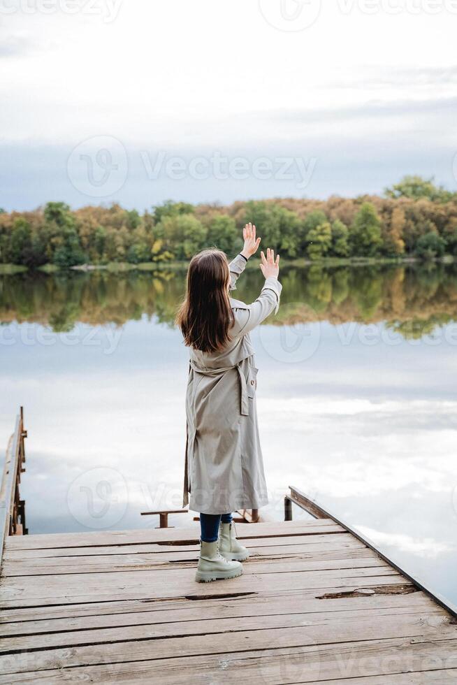 A girl standing in a raincoat on a pyron, a dance of a young beauty in a coat on the bridge, a rear view of a woman in a raincoat, autumn blues, a walk in the park by the lake. photo