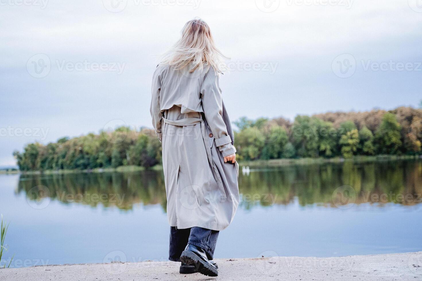 A girl in a raincoat came to the edge of the platform by the water, a gray raincoat, a casual style, a river embankment, a walk by the lake. photo