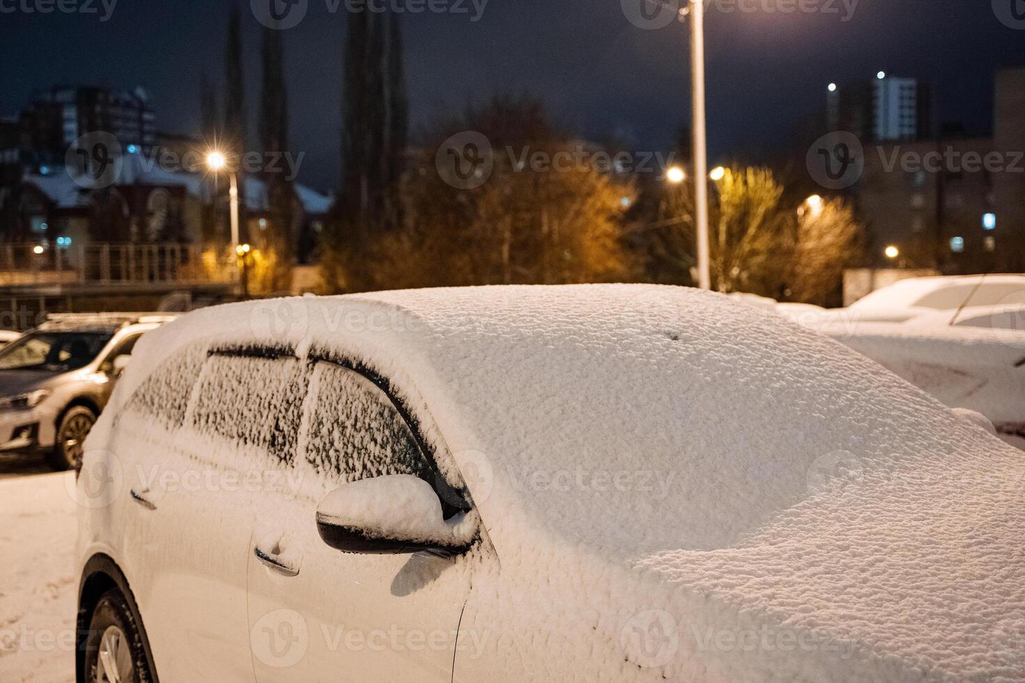 The car is covered with snow. Parking in winter. Winter frosts, street lamp shines on the car. Cleaning the machine from snow photo