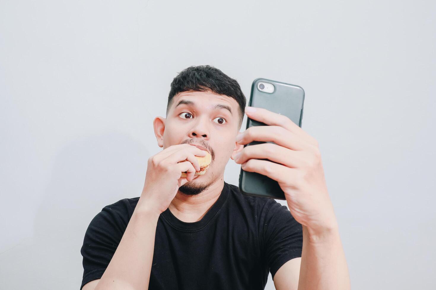 an asian man is enjoying his hamburger while holding his smartphone photo