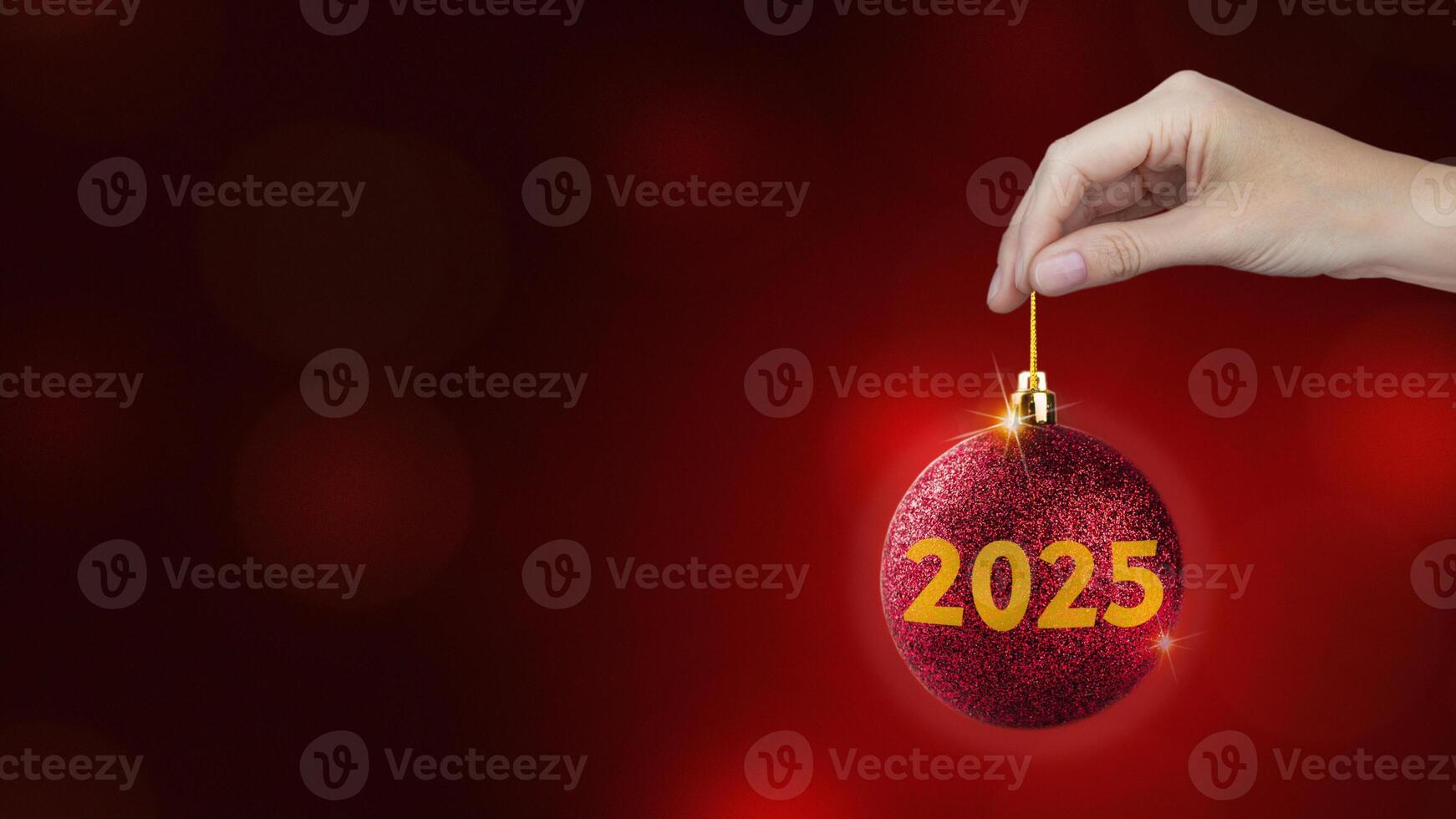 Red Christmas ball with yellow 2025 in woman's hand on red background, New year style copy space photo