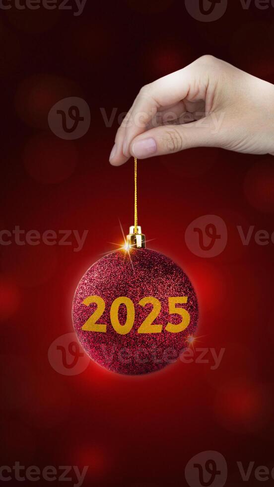 Vertical poster with Red Christmas ball with yellow 2025 in a hand on red background, Xmas, New year style banner photo