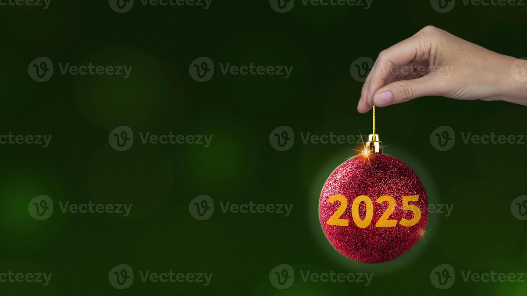 Red Christmas ball with yellow 2025 in woman's hand on green background, horizontal New year style banner copy space photo