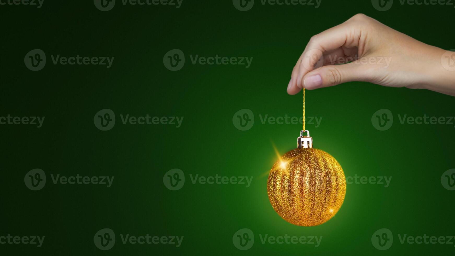Woman's hand holds a Christmas tree golden ball that rotates. New Year, Christmas minimal concept. Green background copy space banner photo