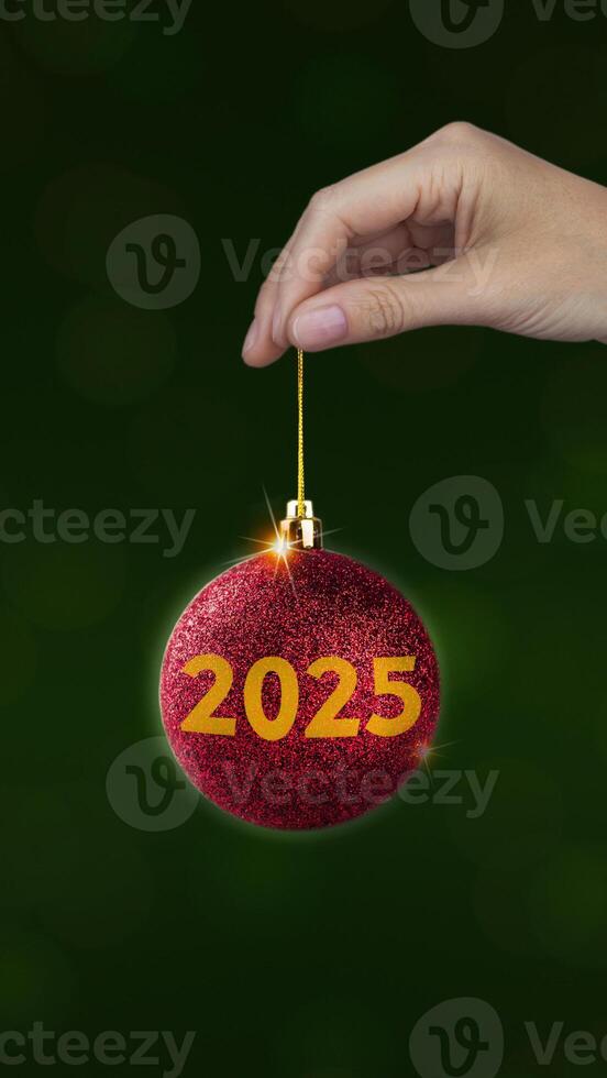 Red Christmas ball with yellow 2025 in woman's hand on green background, vertical New year style photo