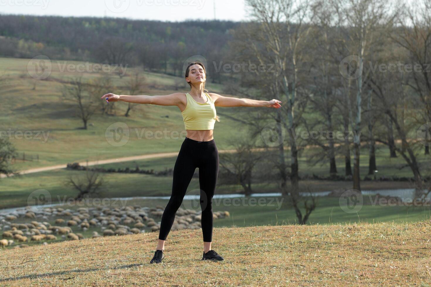 A young slim athletic girl in sportswear performs a set of exercises. Fitness and healthy lifestyle against the background of green spring pasture hills. photo