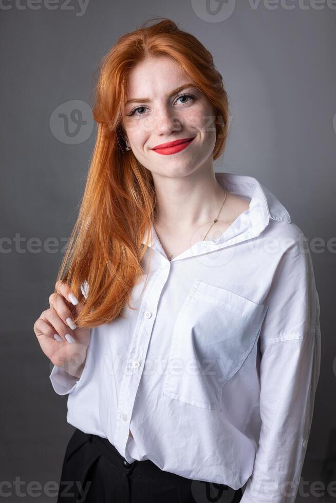 Portrait of a red-haired girl in a white shirt. Woman smiling in studio against gray background. photo