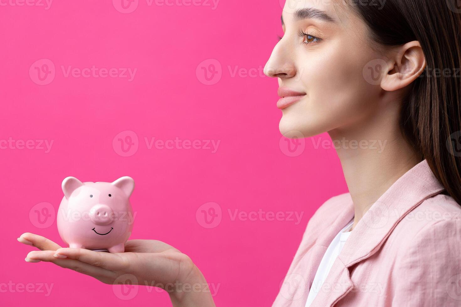 A cute girl in a pink jacket is holding a 3d piggy bank in her hands. Beautiful close-up portrait in studio on pink. photo
