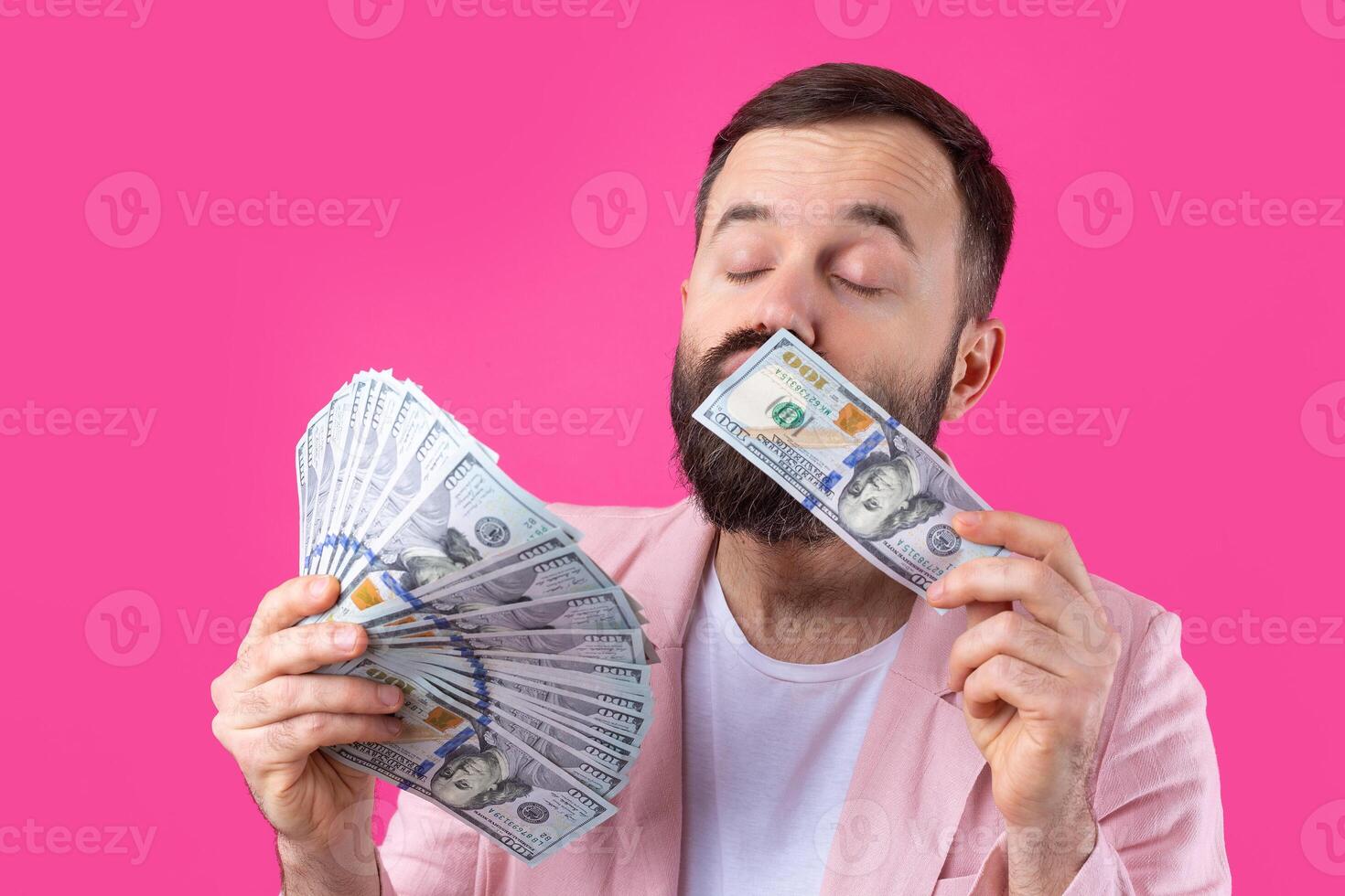 Portrait of a contented young businessman with a beard dressed in a pink jacket showing us dollar banknotes against a red studio background. Taste, smell of money. photo