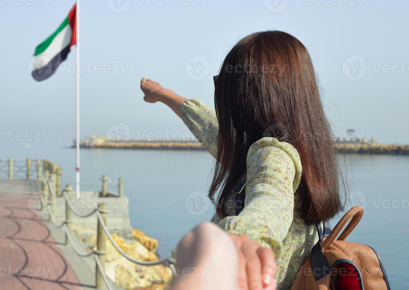 A young woman tourist in a long dress with a backpack points with her hand the direction to the sea to the flag of the United Arab Emirates, and leads her to the pier. photo