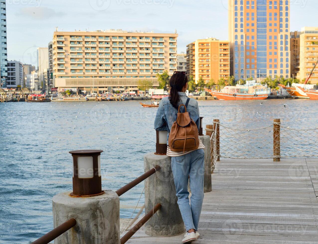 A young woman with a backpack on her back admires the river in the old Dubai Creek. Back view. Journey through the Persian Gulf photo