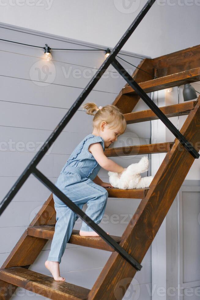 A cute two-year-old girl in a denim jumpsuit with a soft toy in her hands climbs up the stairs to the second floor of the house photo