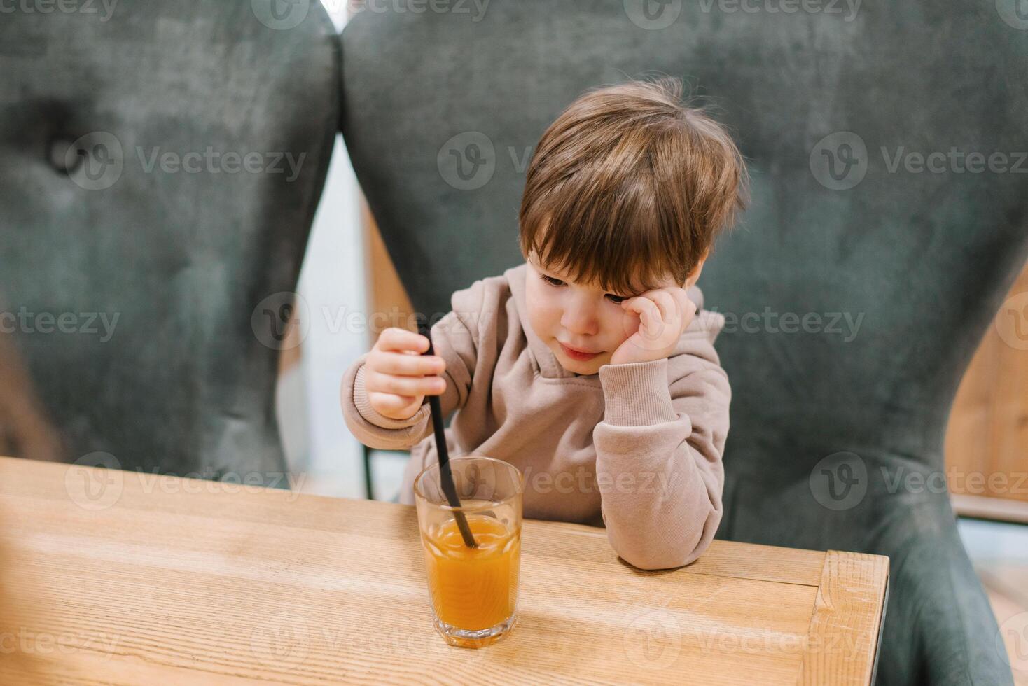 Four year old boy sits at a table in a cafe and looks at a glass of orange juice, holding a straw in his hands photo