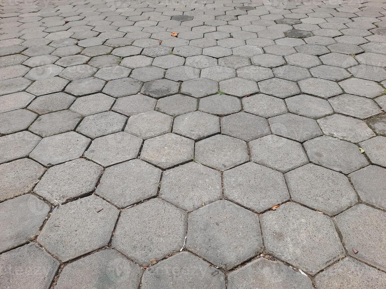 a pavement made of hexagonal concrete tiles arranged in a repeating pattern. The tiles are light gray and have visible wear, with some gaps and small debris between them. photo