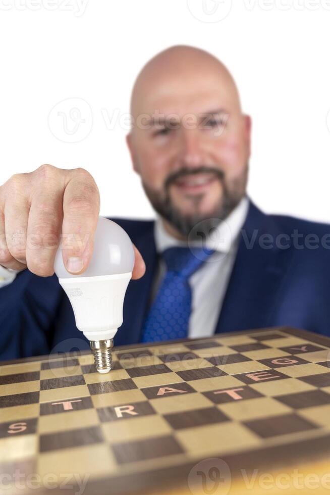 portrait of businessman in blue suit and tie with friendly expression showing light bulb on chessboard as symbol isolated on white background photo