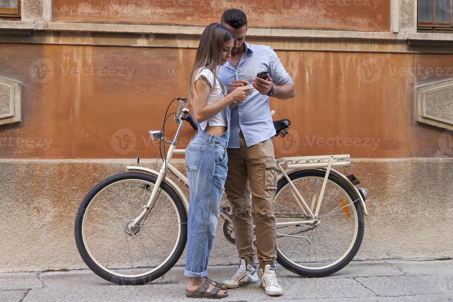 young smiling couple using smartphone while riding bicycles through the streets of a city photo