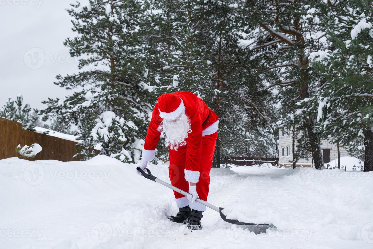Santa Claus cleans snow with shovel in winter outdoors after a snowfall. Cleaning the streets in the village, clearing the passage for cars, difficult weather conditions for Christmas and New Year photo