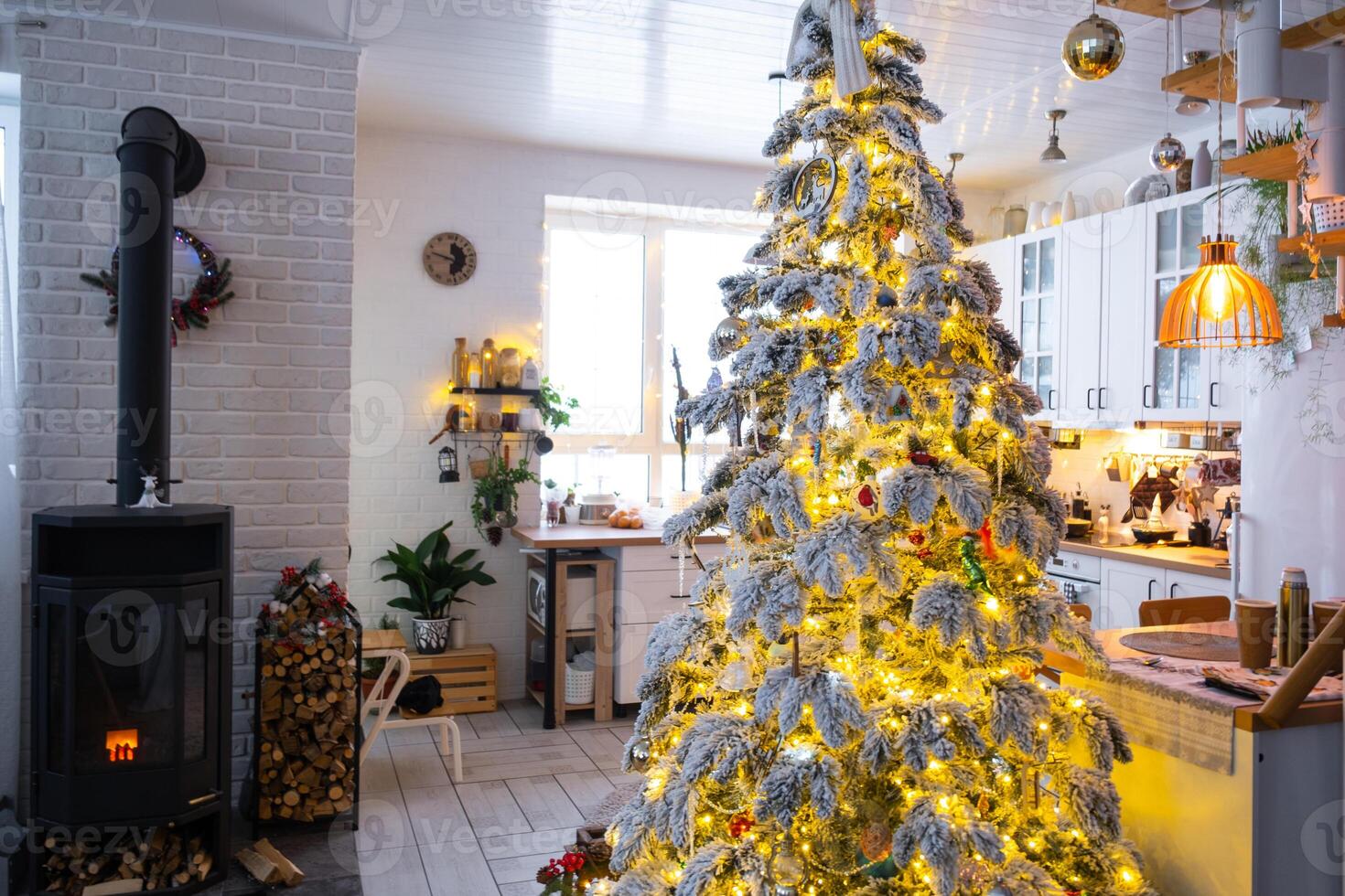 Festive white modern interior of house is decorated for Christmas and New Year in loft style with black stove, fireplace, Christmas tree. Potted plants, firewood in the woodpile photo
