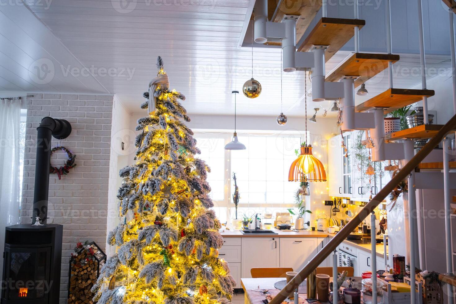 Festive white modern interior of house is decorated for Christmas and New Year in loft style with black stove, fireplace, Christmas tree. Potted plants, firewood in the woodpile photo