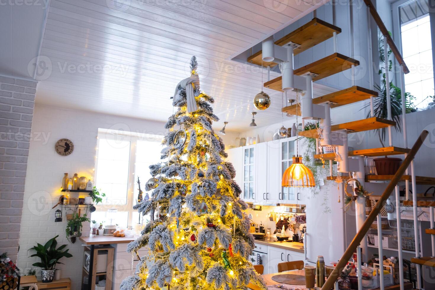 Christmas decor in white kitchen, modern village interior with a snowy Christmas tree and fairy lights. New Year, Christmas mood, cozy home, Metal modular staircase. photo