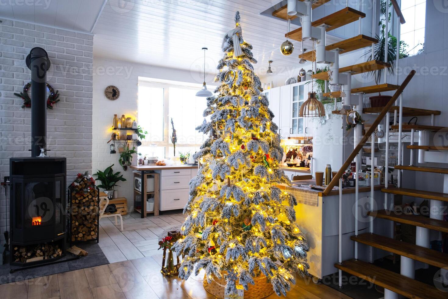Festive white modern interior of house is decorated for Christmas and New Year in loft style with black stove, fireplace, Christmas tree. Potted plants, firewood in the woodpile photo