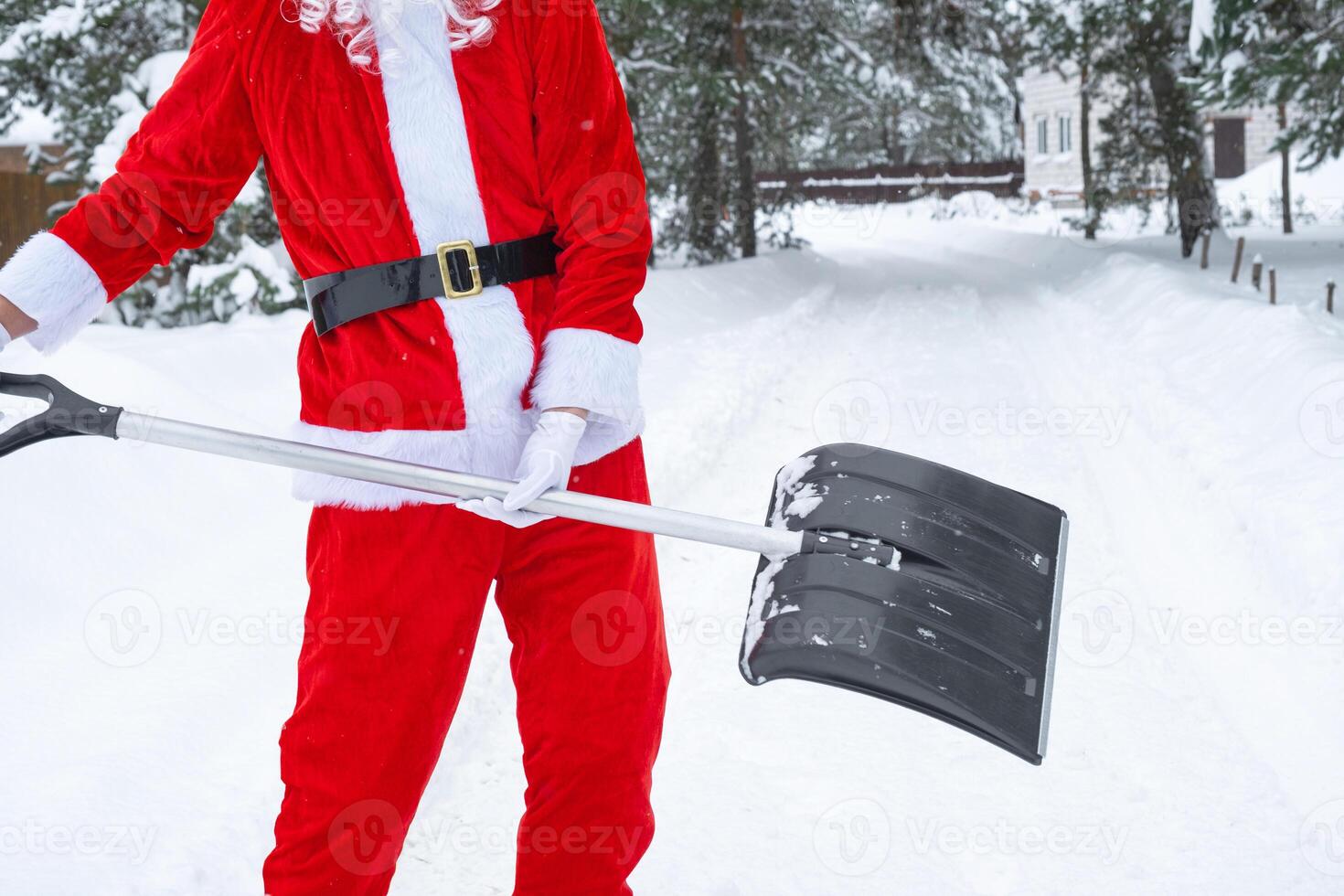 Santa Claus cleans snow with shovel in winter outdoors after a snowfall. Cleaning the streets in the village, clearing the passage for cars, difficult weather conditions for Christmas and New Year photo