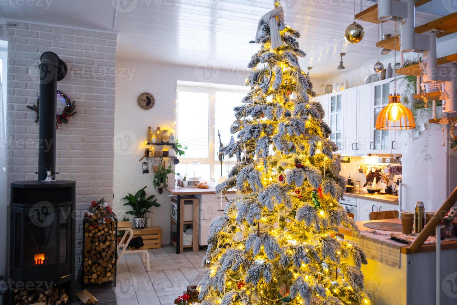 Festive white modern interior of house is decorated for Christmas and New Year in loft style with black stove, fireplace, Christmas tree. Potted plants, firewood in the woodpile photo