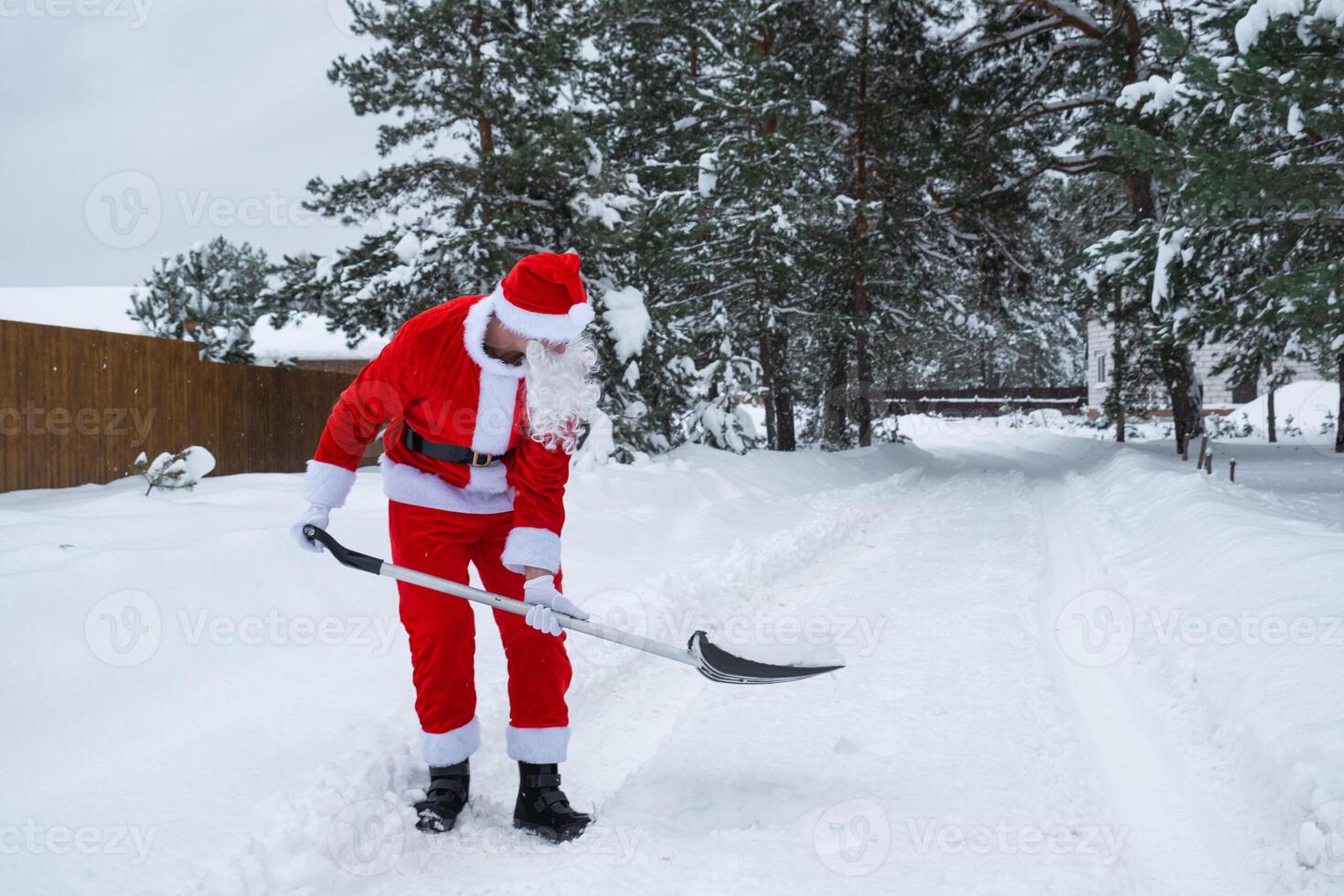 Santa Claus cleans snow with shovel in winter outdoors after a snowfall. Cleaning the streets in the village, clearing the passage for cars, difficult weather conditions for Christmas and New Year photo
