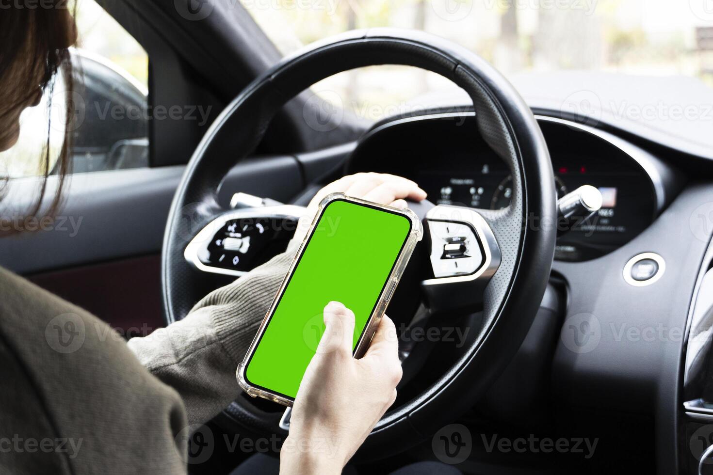 Close-up of woman's hands holding smartphone with green empty copy space on screen mockup behind the wheel of sport's car rear view, connecting phone to a electric car. photo
