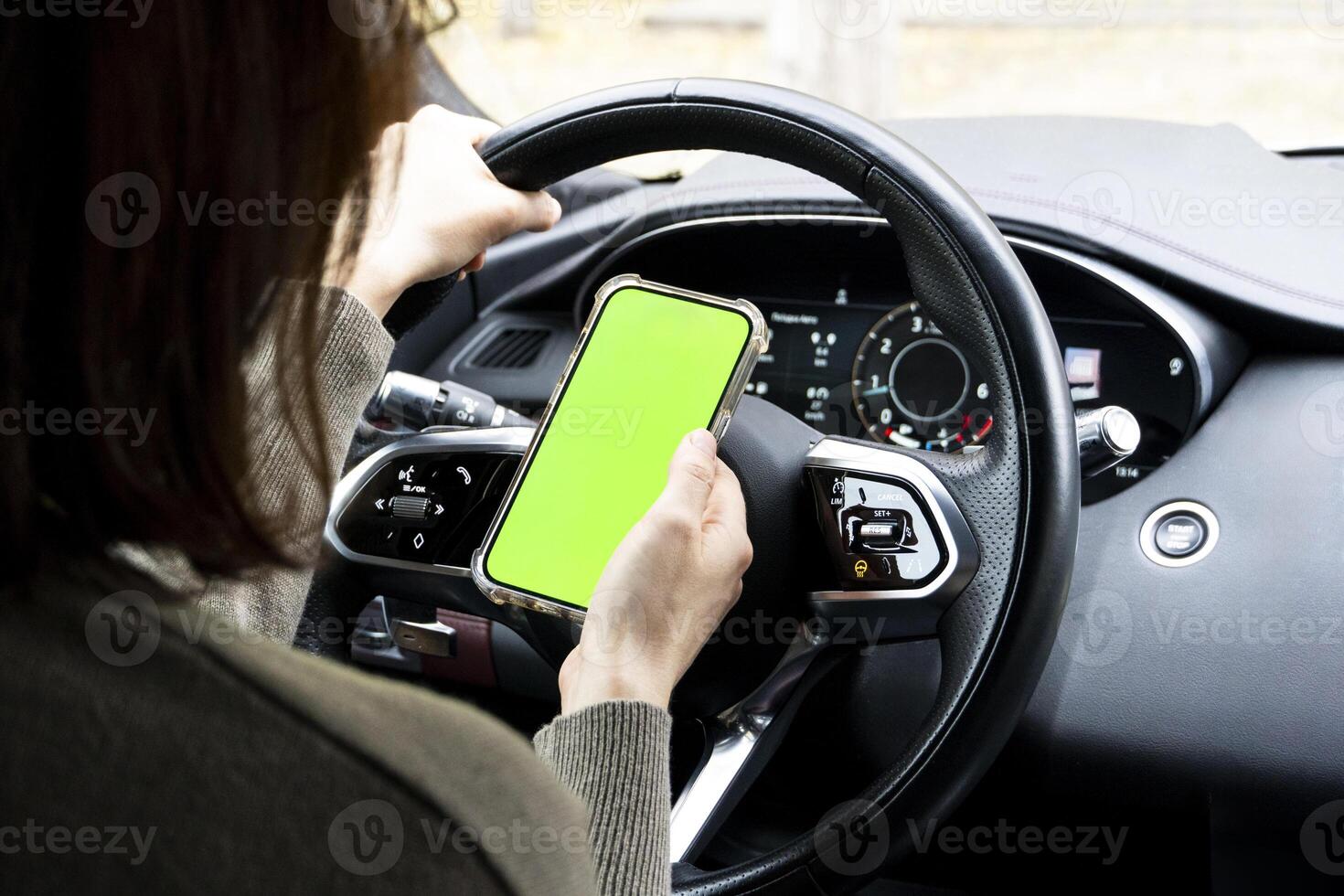 Close-up of woman's hands holding smartphone with green empty copy space on screen mockup behind the wheel of sport's car rear view, connecting phone to a electric car. photo