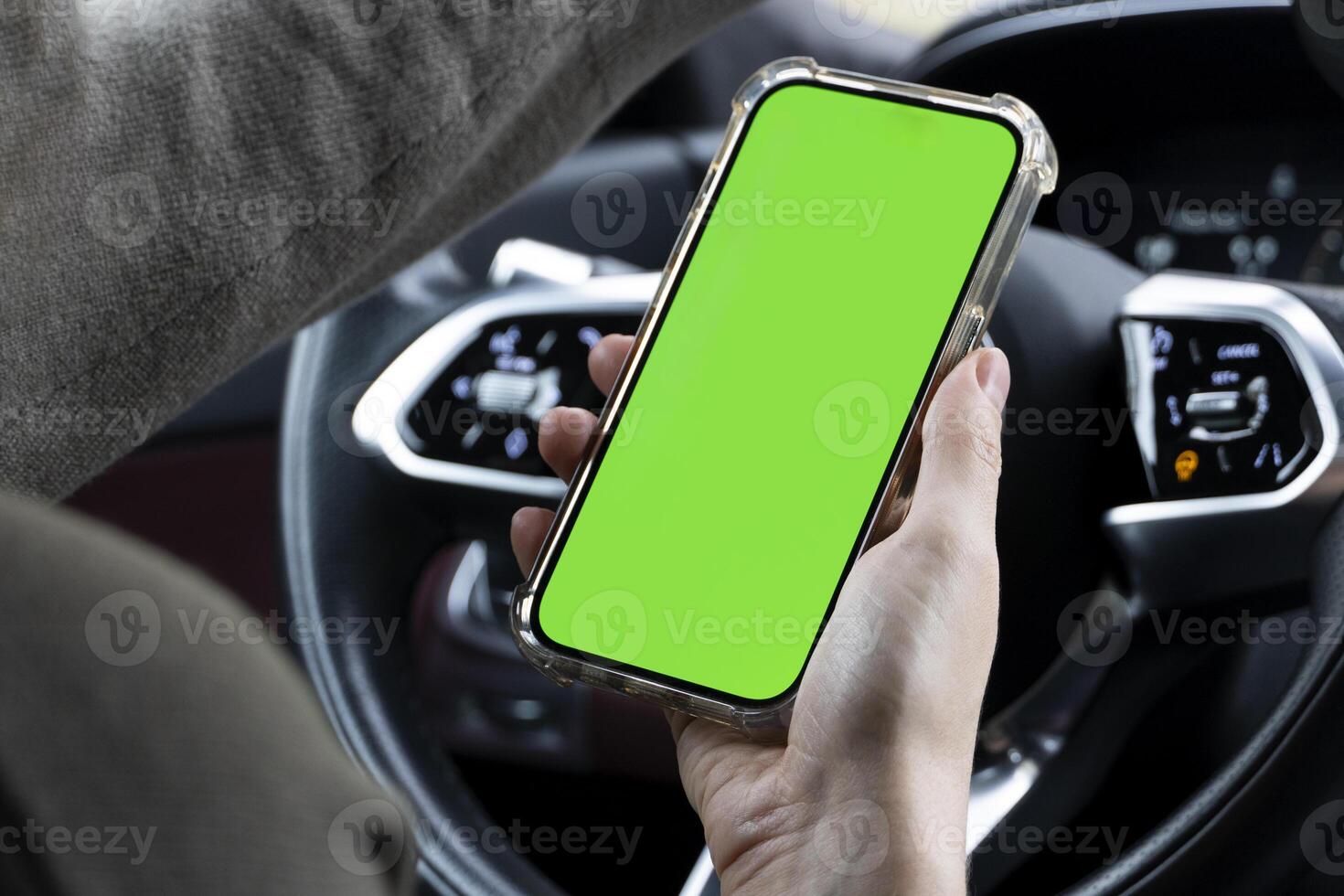 Close-up of woman's hands holding smartphone with green empty copy space on screen mockup behind the wheel of sport's car rear view, connecting phone to a electric car. photo