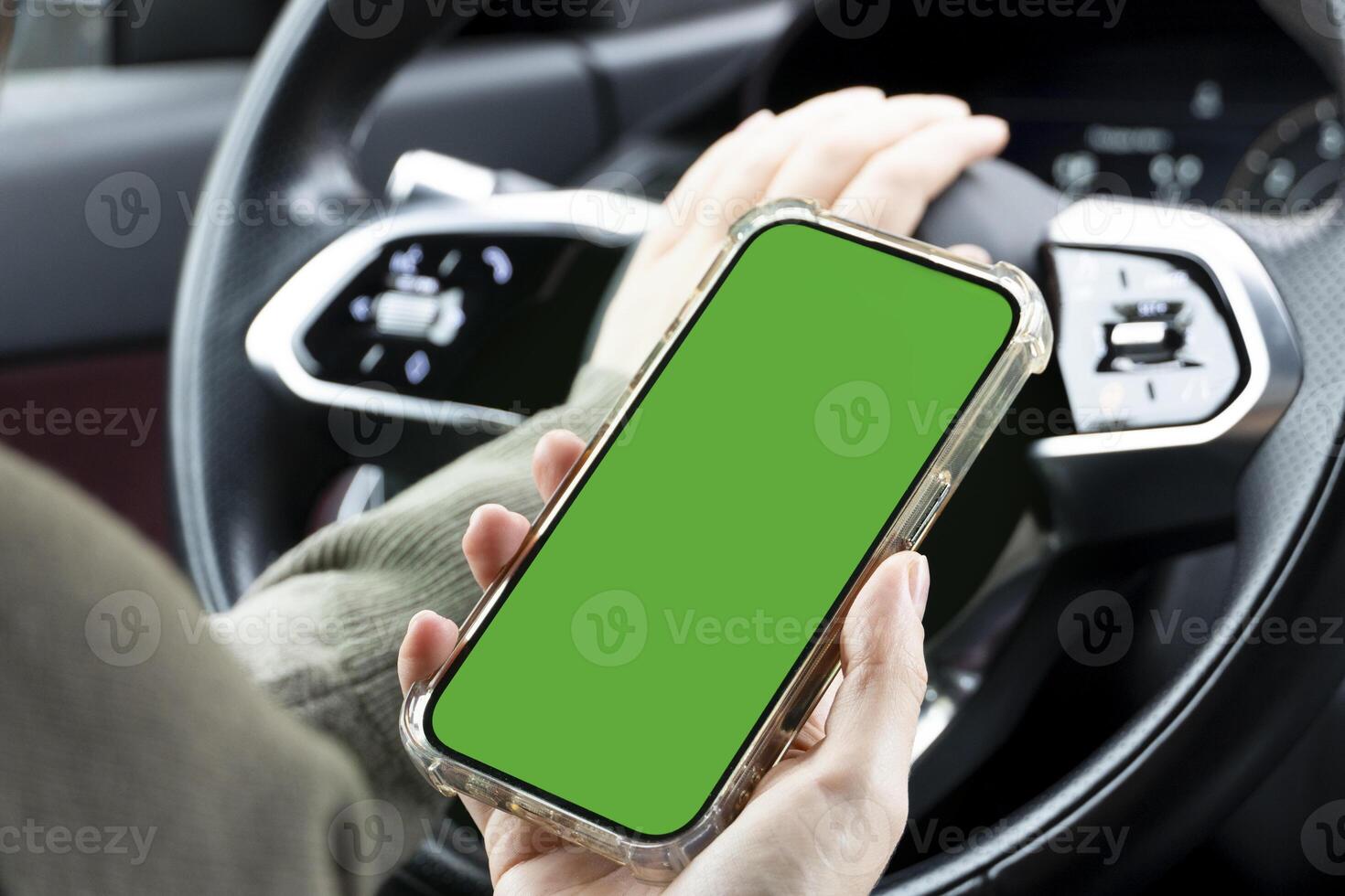 Close-up of woman's hands holding smartphone with green empty copy space on screen mockup behind the wheel of sport's car rear view, connecting phone to a electric car. photo