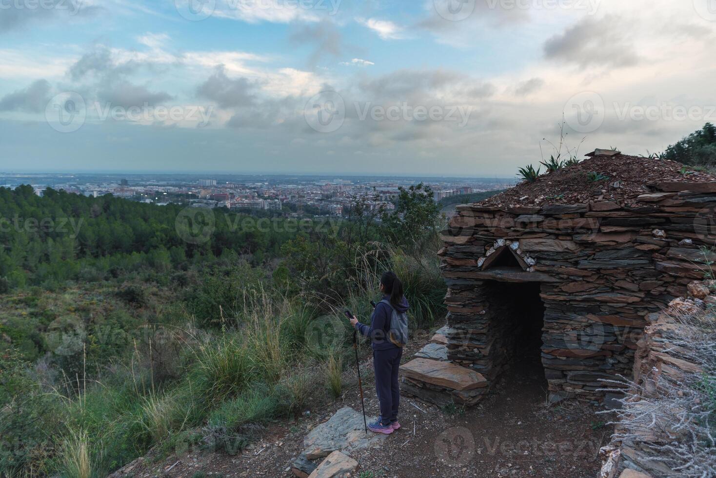 A hiker standing next to a stone shelter contemplates the mountains, city and sea in the background. photo