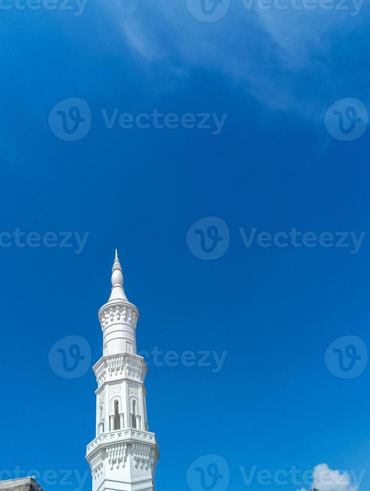 A pristine white minaret, intricately designed, stands tall against a backdrop of a clear, deep blue sky. photo