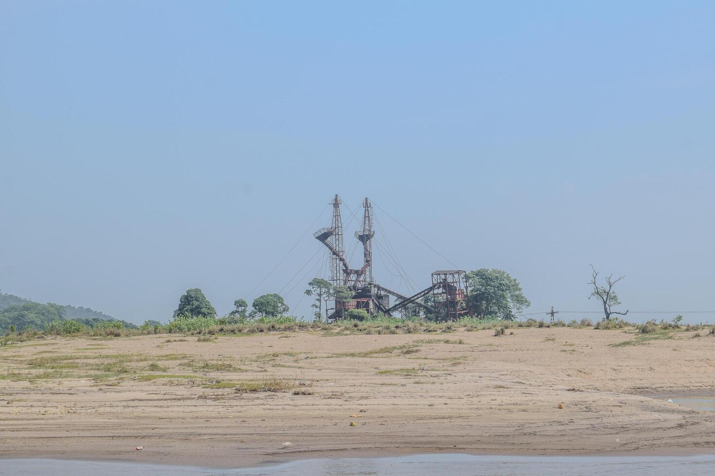 An industrial facility stands prominently on the sandy riverbank, surrounded by sparse greenery and mountains on a sunny day, highlighting the contrast between nature and industry. photo