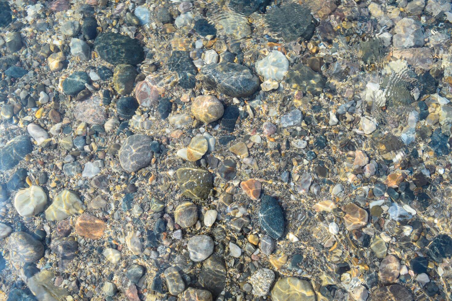 The sunlight shines through clear water, showcasing an array of colorful stones and pebbles on a natural lakeside. This tranquil setting invites relaxation and exploration. photo