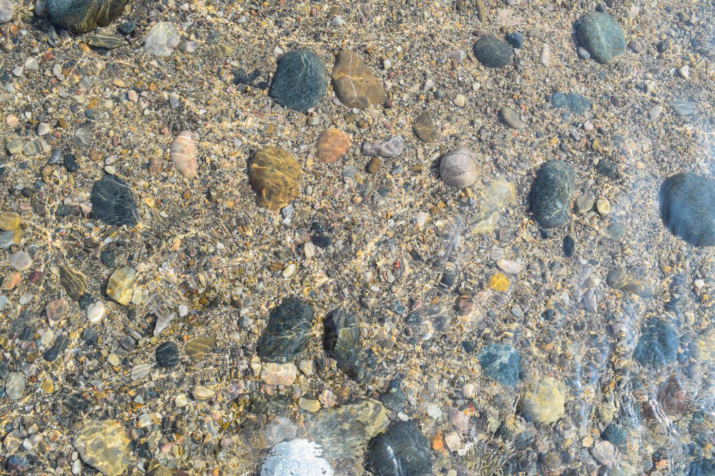 The calm surface of a lake showcases a variety of colorful pebbles submerged beneath clear water, reflecting sunlight on a beautiful day. photo