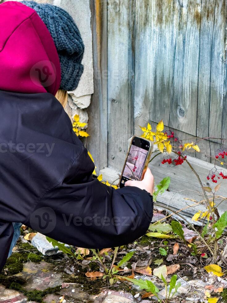 A young woman crouches near an old, weathered wooden door, capturing the details of bright red autumn berries and yellow leaves with her smartphone. Dressed in a dark jacket and burgundy hoodie, she photo
